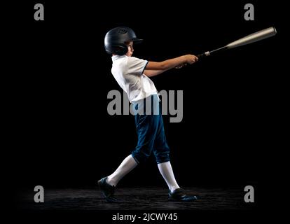 Ritratto dinamico di piccolo giocatore di baseball, caraffa in uniforme blu-bianco formazione isolato su sfondo nero studio. Concetto di sport, realizzazioni Foto Stock