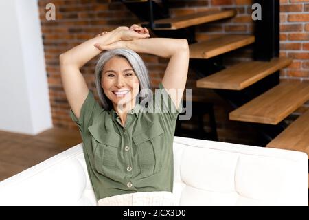 Donna di mezza età positiva con capelli lunghi al divano in appartamento moderno. 50s femmina asiatica che riposa a casa, guarda la macchina fotografica. Concetto di benessere Foto Stock