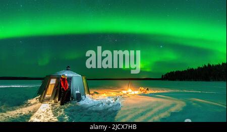 Escursionista che guarda l'aurora boreale (aurora boreale) vicino al falò e igloo nel paesaggio ghiacciato, Jokkmokk, Lapponia, Svezia, Scandinavia Foto Stock