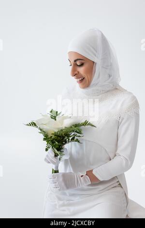 felice sposa musulmana in guanti e abito da sposa che tiene calla giglio fiori isolato su grigio Foto Stock