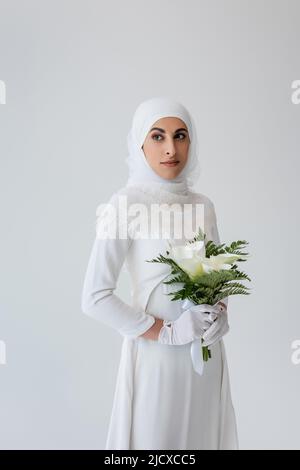 sposa musulmana in guanti e abito da sposa che tiene bouquet di calla giglio isolato sul grigio Foto Stock