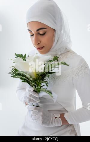 sposa musulmana in guanti e abito da sposa che puzzano fiori di calla giglio isolato su grigio Foto Stock