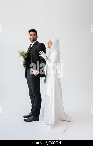 felice sposa musulmana in hijab rugging sposo con bouquet su grigio Foto Stock