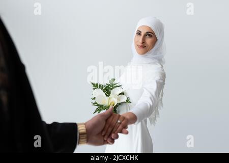 felice sposa musulmana con bouquet che tiene le mani con lo sposo isolato sul grigio Foto Stock