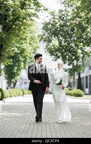 buon sposo che tiene le mani con sposa musulmana con bouquet di nozze e camminare insieme Foto Stock