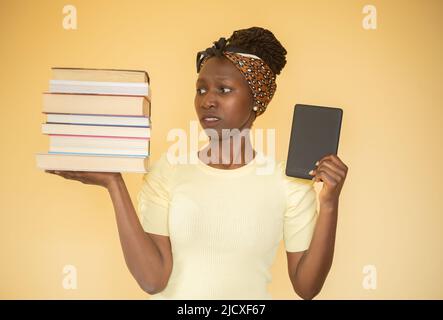 giovane donna che confronta libri stampati con e-book mentre accigliava a libri sulla sua mano destra e e-book sulla sua mano sinistra Foto Stock