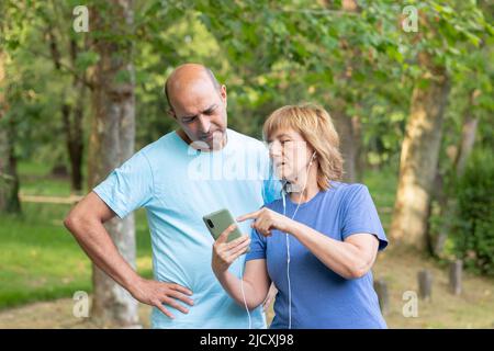 Due anziani eterosessuali che guardano verso il basso e verso il mobile mentre si riposano facendo sport nel parco naturale Foto Stock