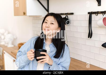 Una donna asiatica attraente riposa in cucina, tiene una tazza con una bevanda calda e guarda nella macchina fotografica. Foto Stock