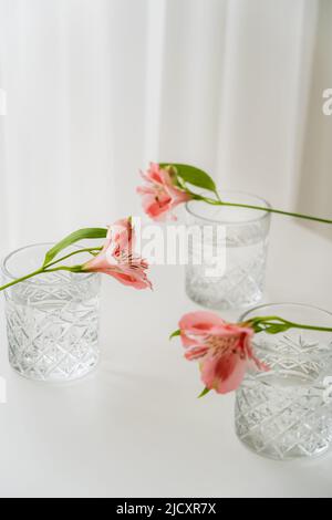 vetri di cristallo con acqua vicino ai fiori di alstroemeria su piano bianco e sfondo grigio Foto Stock