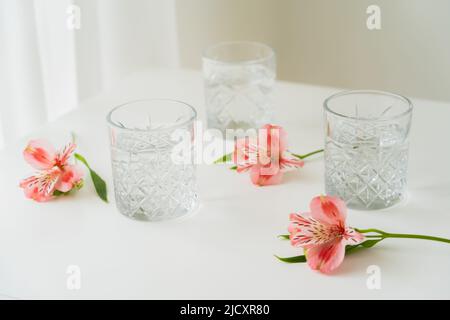 vetri di cristallo con acqua vicino ai fiori rosa su superficie bianca e sfondo grigio Foto Stock