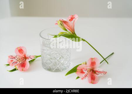 fiori di alstroemeria vicino vetro sfaccettato con acqua pulita su superficie bianca e sfondo grigio Foto Stock