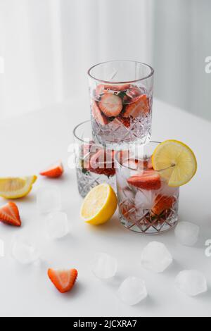 bicchieri con tonico freddo alla fragola vicino ai limoni tagliati e cubetti di ghiaccio sul piano portapaziente bianco Foto Stock