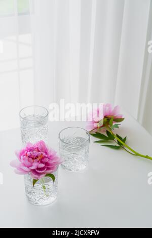 occhiali trasparenti e peonie rosa su un tavolo bianco vicino alla tenda Foto Stock