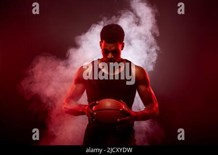 Lato giocatore di basket illuminato con colore rosso che tiene una palla contro sfondo fumo. Uomo africano americano concentrato serio. Foto Stock