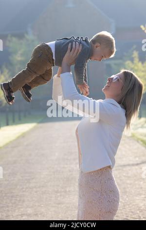 una giovane e felice madre e un bambino, giocando, si erge nel mezzo di un parco ai raggi del sole mattutino nascente. Foto di alta qualità Foto Stock