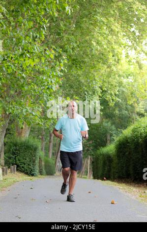 Uomo acconciato maturo in abbigliamento sportivo che corre lungo un percorso in cemento tra i cespugli e gli alberi della natura Foto Stock