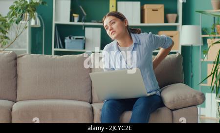 Freelancer seduto sul divano lavoro a distanza da casa improvvisamente sensazione di dolore alla schiena e alla vita. Donna sensazione stanca di lungo sedentary Lifestyle provare Foto Stock