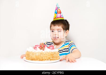 Un bambino in un cappellino festivo guarda una torta con una candela nella forma del numero tre. Foto Stock