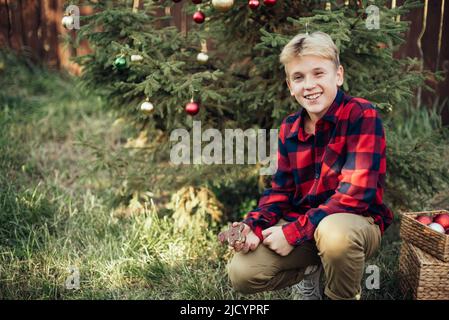 Natale nel mese di luglio. Bambino in attesa di Natale in legno in estate. Ritratto di ragazzo in abito rosso decorando albero di natale. Vacanze invernali e concetto di gente. Buon Natale e buone feste Foto Stock