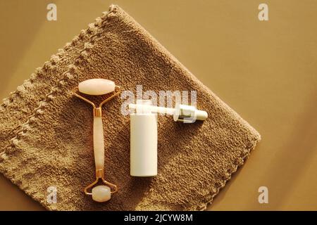 Set per la cura del viso a casa su un asciugamano marrone. Rullo per massaggio viso al quarzo rosa o gua sha e siero in una bottiglia di vetro bianco e pipetta per la cura del viso. Foto Stock