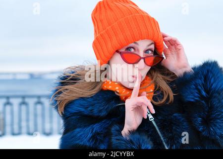 Primo piano Portrait Funny Girl in Orange Beanie Hat e Blue Fur Coat su sfondo neve invernale. Mysterious Young Woman indossa eleganti occhiali da sole arancioni Foto Stock