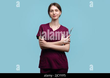 Giovane chirurgo femminile in posa con strumenti chirurgici in acciaio su sfondo blu studio. Donna medico dipendente medico indossa Medical uniforme chirurgico su Foto Stock