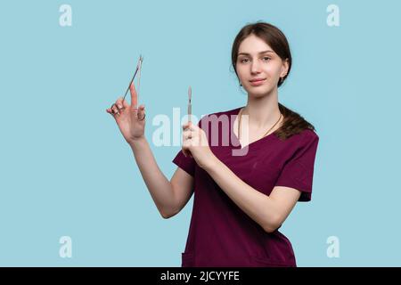 Giovane chirurgo femminile in posa con strumenti chirurgici in acciaio su sfondo blu studio. Donna medico dipendente medico indossa Medical uniforme chirurgico su Foto Stock