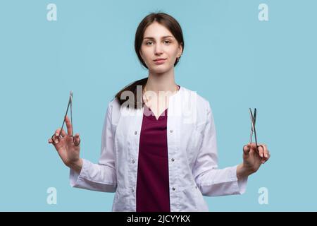 Giovane chirurgo femminile in posa con strumenti chirurgici in acciaio su sfondo blu studio. Donna medico dipendente medico indossa uniforme medica mantello bianco. Foto Stock