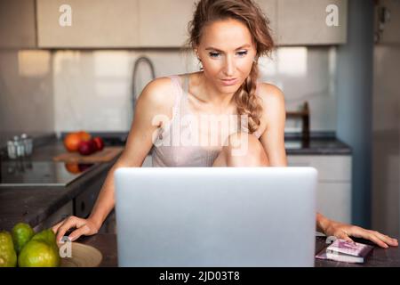 Una giovane bella donna di buon umore comunica via video link su un computer portatile in cucina della casa in prima serata al tramonto. Foto Stock