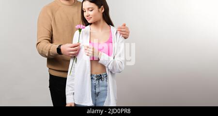 bella giovane donna in abiti moderni prendendo un fiore dal ragazzo in piedi dietro di lei e abbracciarla a spalla. Foto Stock