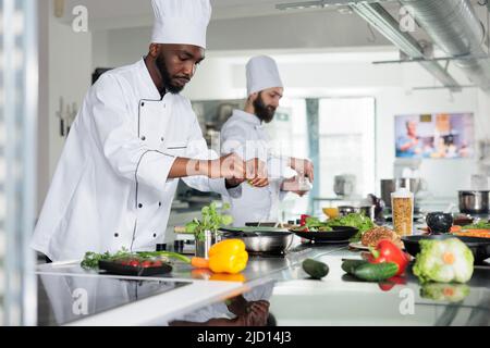 Esperto di gastronomia che indossa uniforme di cucina mentre prepara deliziosi piatti gourmet per il servizio cena al ristorante. Capo chef che ha padella su cucina a gas mentre cucinando gustoso cibo pasto in cucina. Foto Stock