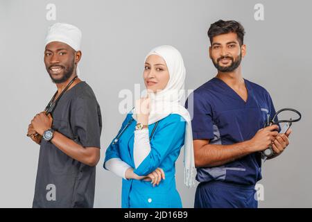 Gruppo di paramedici sorridenti su sfondo grigio. donna medico musulmana con colleghi chirurgo africano e indiano Foto Stock