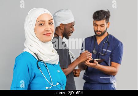 Gruppo di paramedici sorridenti su sfondo grigio. donna medico musulmana con colleghi chirurgo africano e indiano Foto Stock