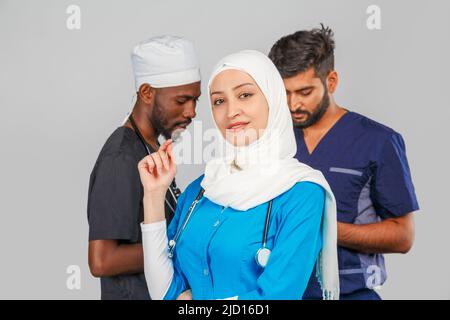 Gruppo di paramedici sorridenti su sfondo grigio. donna medico musulmana con colleghi chirurgo africano e indiano Foto Stock