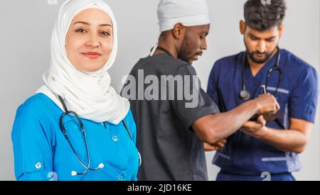 Gruppo di paramedici sorridenti su sfondo grigio. donna medico musulmana con colleghi chirurgo africano e indiano Foto Stock