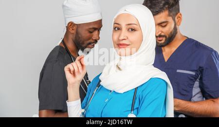 Gruppo di paramedici sorridenti su sfondo grigio. donna medico musulmana con colleghi chirurgo africano e indiano Foto Stock