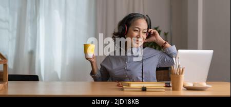 Donna asiatica felice e anziana che indossa le cuffie ascolta le canzoni sul laptop in salotto in casa accogliente Foto Stock