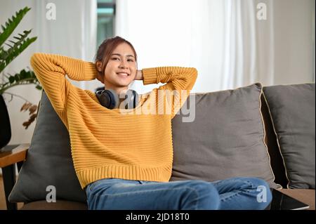 Felice giovane donna asiatica millenaria con cuffie rilassa chilling nel soggiorno, seduta e appoggiata sul divano. Foto Stock