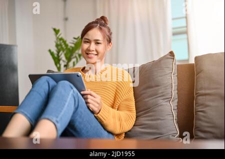 Un'attraente donna asiatica millenaria si rilassa nel soggiorno, si siede su un comodo divano e utilizza un touchpad portatile tablet. Foto Stock