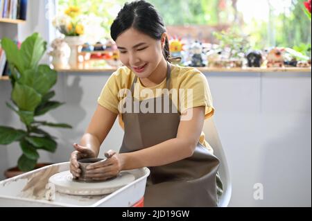 Bella e affascinante giovane donna asiatica che fa una tazza di ceramica sulla ruota di un vasaio nel laboratorio artigianale. Foto Stock