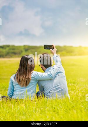 Persone innamorate che prendono selfie sul campo con il loro smartphone, sorridente coppia innamorata seduta sull'erba che prende selfie, giovane coppia innamorata Foto Stock