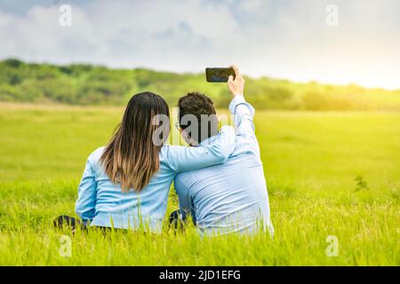 Giovane coppia innamorata che prende un selfie sul campo, gente innamorata che prende selfie sul campo con il loro smartphone, sorridente coppia innamorata seduta Foto Stock