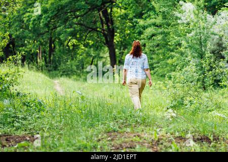 Ritratto di giovane donna buxom con lunghi capelli rossi ricci che camminano nella foresta del parco tra alberi verdi cespugli. Estate, natura. Foto Stock