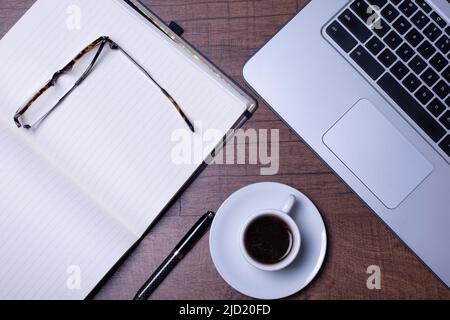 Tavolo in legno con computer, caffè espresso in una tazza bianca su una pentola bianca, bicchieri, penna e notebook. Foto Stock
