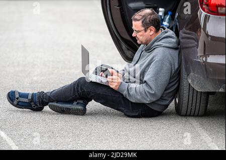 uomo di mezza età seduto sull'asfalto vicino all'auto e lavorando su un computer portatile, lavorando a distanza, viaggiare Foto Stock