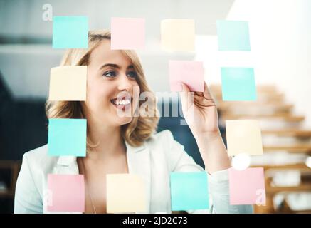 Generazione di idee geniali. Scatto di una donna d'affari che ha organizzato note appiccicose su una parete di vetro in un ufficio moderno. Foto Stock