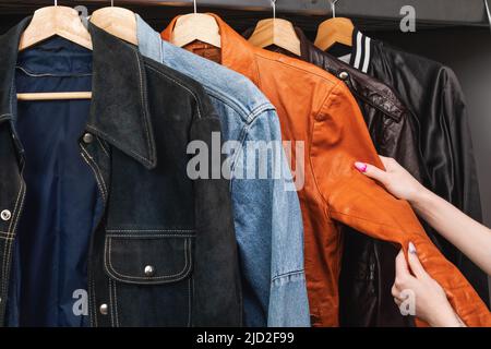 Donna che sceglie l'abbigliamento in un negozio di seconda mano. Vari giacche vintage in pelle scamosciata e jeans appendono sul portabiti. Thrifting e sostenibilità in Foto Stock