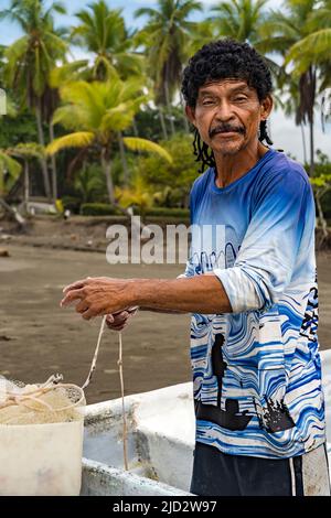 Uomo che guarda la macchina fotografica che prepara la sua rete a pescare nella sua barca con palme sullo sfondo. Foto Stock