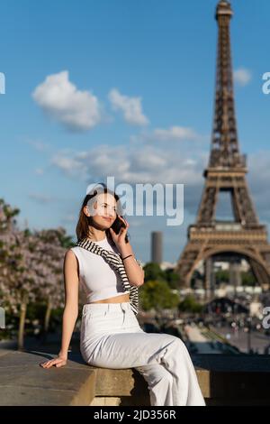 giovane donna sorridente in abito elegante che parla sullo smartphone mentre si siede vicino alla torre eiffel di parigi Foto Stock