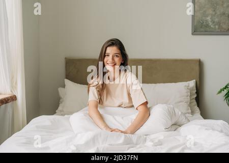 Giovane sorridente riposava donna di bruna latina seduta felicemente sul letto in una camera da letto accogliente e luminosa. Foto Stock